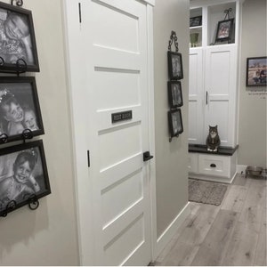 Pu&ograve; includere: Una porta bianca con un cartello che recita "Rest Room" e una maniglia della porta nera. Ci sono foto incorniciate sul muro a sinistra della porta e un gatto seduto su una panca a destra della porta.