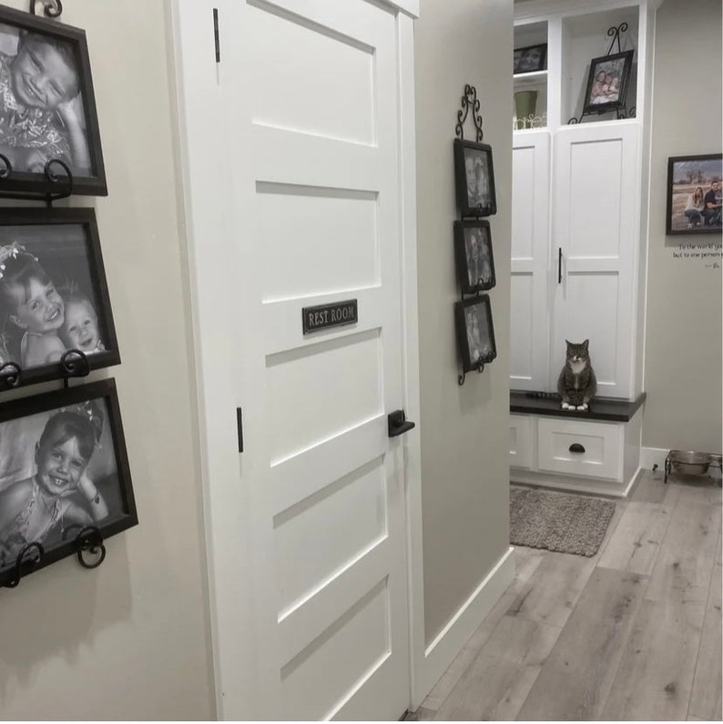 Pu&ograve; includere: Una porta bianca con un cartello che recita "Rest Room" e una maniglia della porta nera. Ci sono foto incorniciate sul muro a sinistra della porta e un gatto seduto su una panca a destra della porta.
