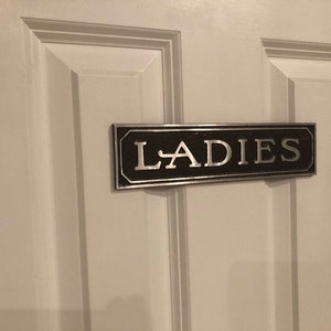 K&ouml;nnte beinhalten: Ein schwarzes und silbernes Schild mit dem Wort "LADIES" in silbernen Buchstaben auf einer wei&szlig;en T&uuml;r.