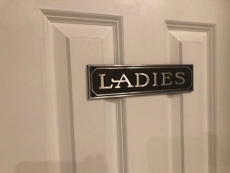 K&ouml;nnte beinhalten: Ein schwarzes und silbernes Schild mit dem Wort "LADIES" in silbernen Buchstaben auf einer wei&szlig;en T&uuml;r.