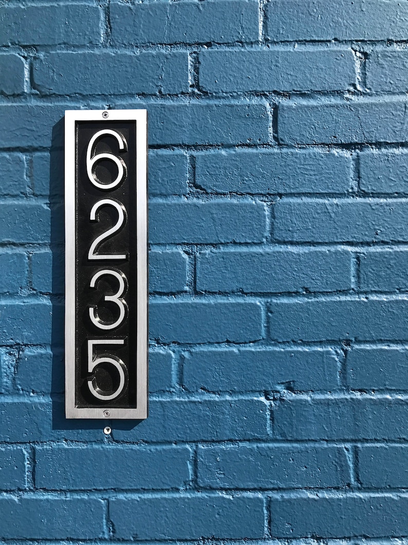 May include: A silver metal house number plaque with black numbers reading "6235" mounted on a blue brick wall.