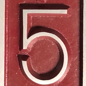 May include: A close-up of a red number five with a white outline. The number is raised and has a textured surface.
