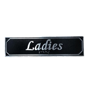 May include: Black rectangular sign with silver lettering that reads "Ladies" with braille below.