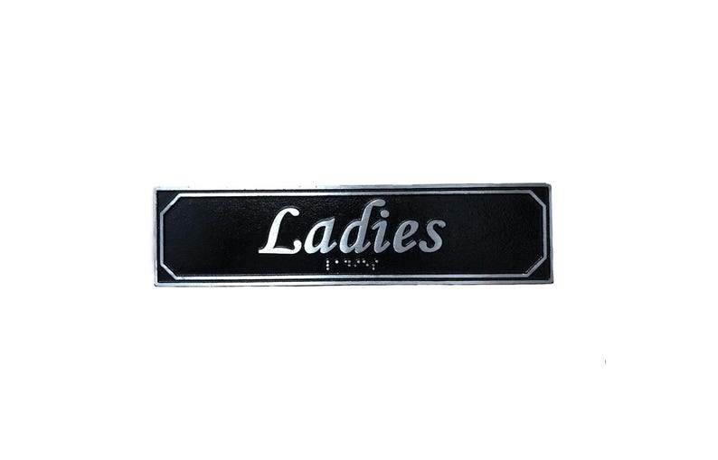 May include: Black rectangular sign with silver lettering that reads "Ladies" with braille below.