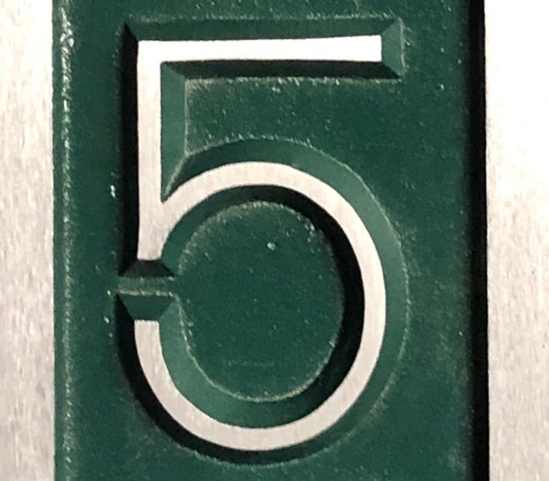 May include: A close-up of a green metal number five with a white outline. The number is raised and has a three-dimensional appearance.