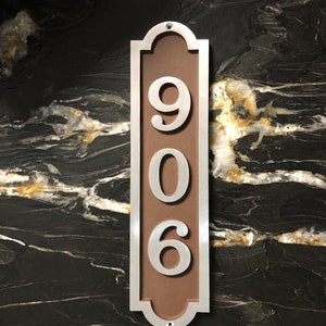 May include: A silver metal house number plaque with a brown background. The number 906 is displayed in white on the plaque.