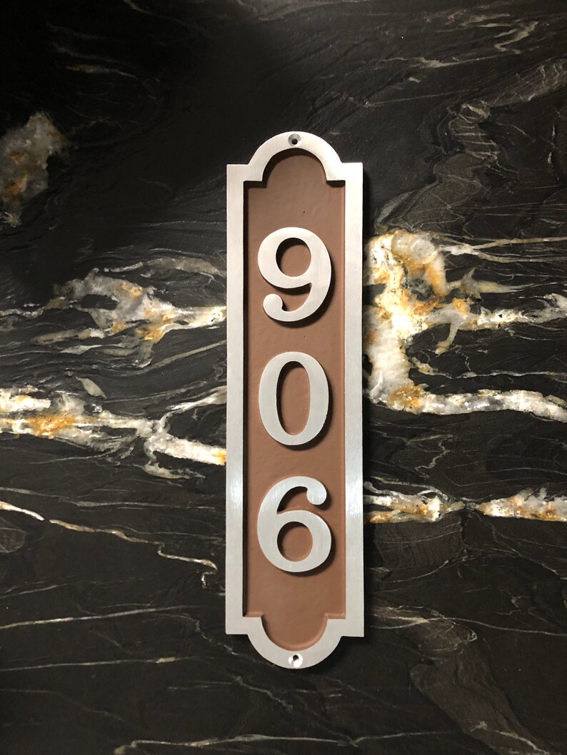 May include: A silver metal house number plaque with a brown background. The number 906 is displayed in white on the plaque.