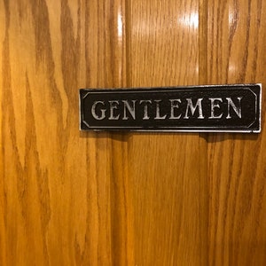 May include: A black and silver sign with the word "GENTLEMEN" in white lettering.