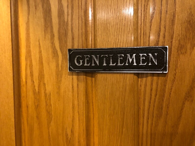 May include: A black and silver sign with the word "GENTLEMEN" in white lettering.