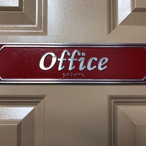 May include: A red rectangular sign with a silver border and the word "Office" in silver lettering. The sign has braille lettering below the word "Office".