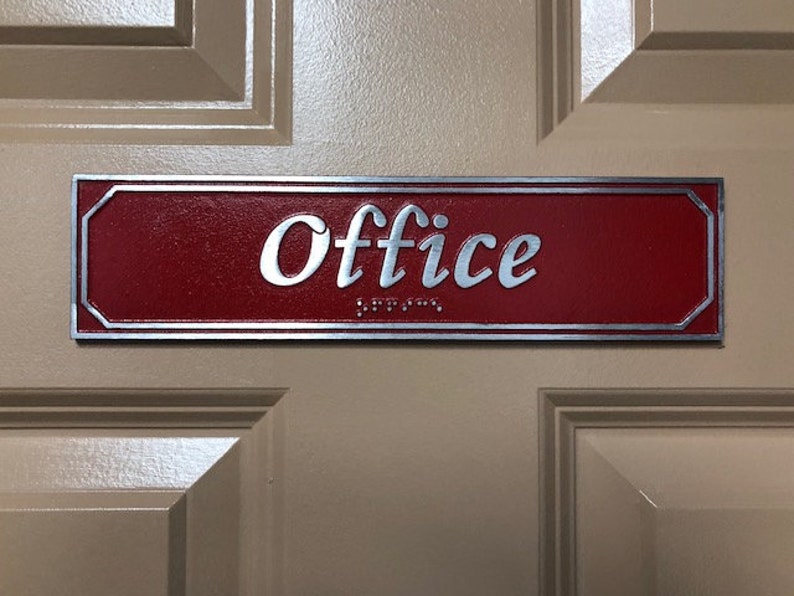 May include: A red rectangular sign with a silver border and the word "Office" in silver lettering. The sign has braille lettering below the word "Office".