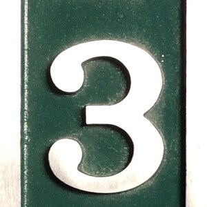 Puede incluir: Un n&uacute;mero tres blanco sobre un fondo verde oscuro. El n&uacute;mero est&aacute; recortado del fondo y est&aacute; en relieve.