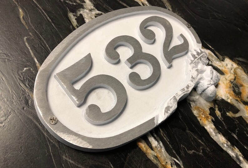 May include: A white oval-shaped house number sign with silver raised numbers "532" and a decorative acorn on the side.