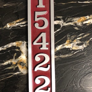 May include: A vertical, oval-shaped house number sign with the numbers 15422 in silver on a burgundy background.