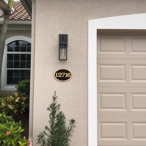 May include: A black oval house number sign with gold lettering, reading "12716", is mounted on a beige stucco wall. A black outdoor light fixture is mounted above the house number sign.