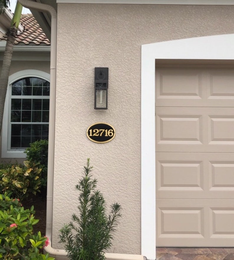 May include: A black oval house number sign with gold lettering, reading "12716", is mounted on a beige stucco wall. A black outdoor light fixture is mounted above the house number sign.