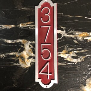 May include: A vertical house number sign with the numbers 3754 in white on a red background. The sign is made of metal and has a silver border.