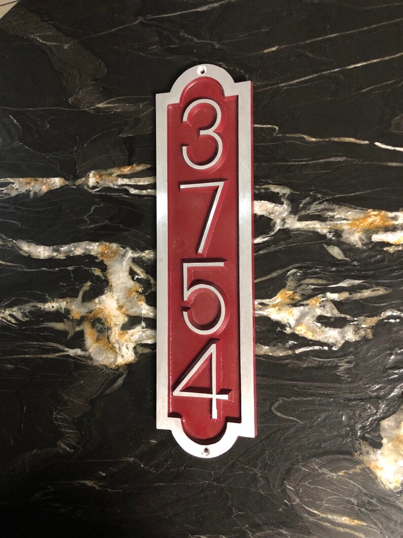 May include: A vertical house number sign with the numbers 3754 in white on a red background. The sign is made of metal and has a silver border.