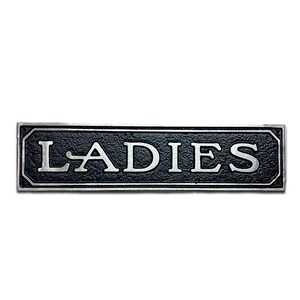 K&ouml;nnte beinhalten: Ein schwarzes und silbernes Metallschild mit dem Wort "LADIES" in wei&szlig;en Buchstaben.