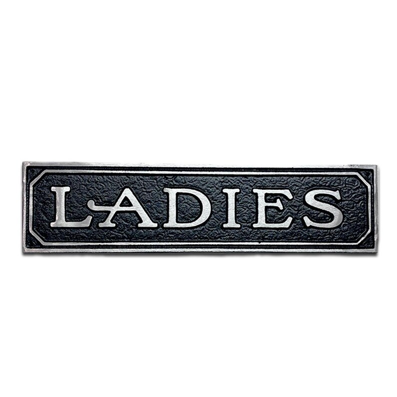 K&ouml;nnte beinhalten: Ein schwarzes und silbernes Metallschild mit dem Wort "LADIES" in wei&szlig;en Buchstaben.