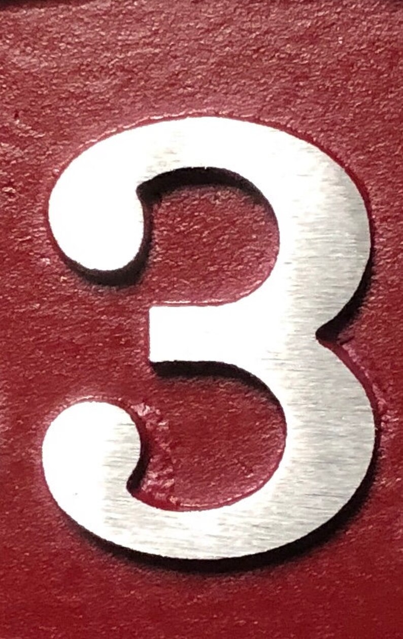 May include: A close-up of the number 3, crafted from brushed metal, set against a deep red background. The number has a classic, rounded font, with a slightly textured surface. The background has a textured, painted appearance.