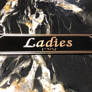 May include: A black rectangular sign with gold trim and the word "Ladies" in gold lettering. The sign also includes braille lettering.