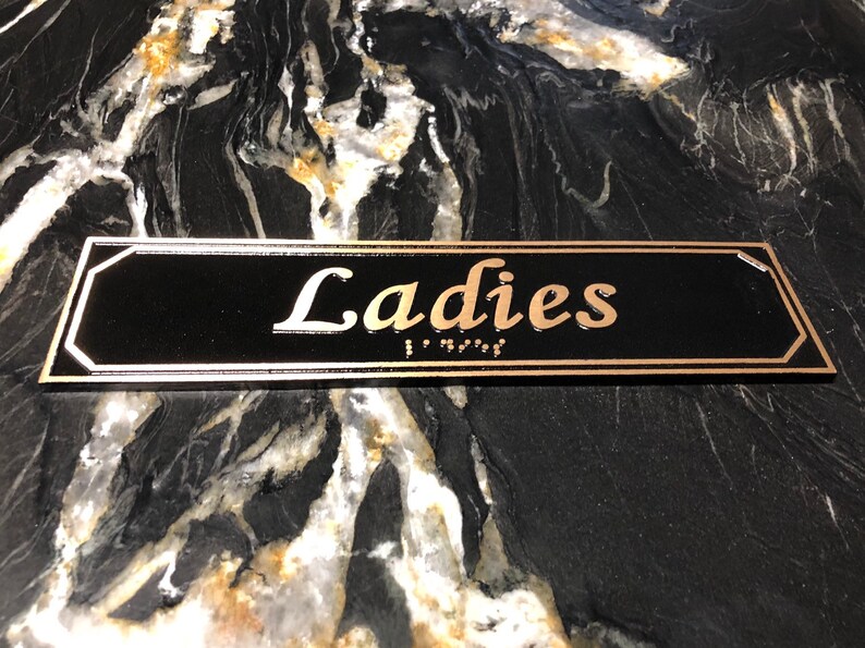May include: A black rectangular sign with gold trim and the word "Ladies" in gold lettering. The sign also includes braille lettering.