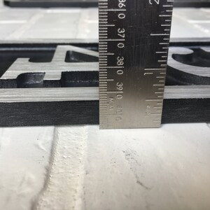 Puede incluir: Primer plano de una placa de n&uacute;mero de casa de metal negro y plateado con el n&uacute;mero "4" en ella. Una regla se coloca junto a la placa para mostrar el tama&ntilde;o. La regla est&aacute; marcada en pulgadas.