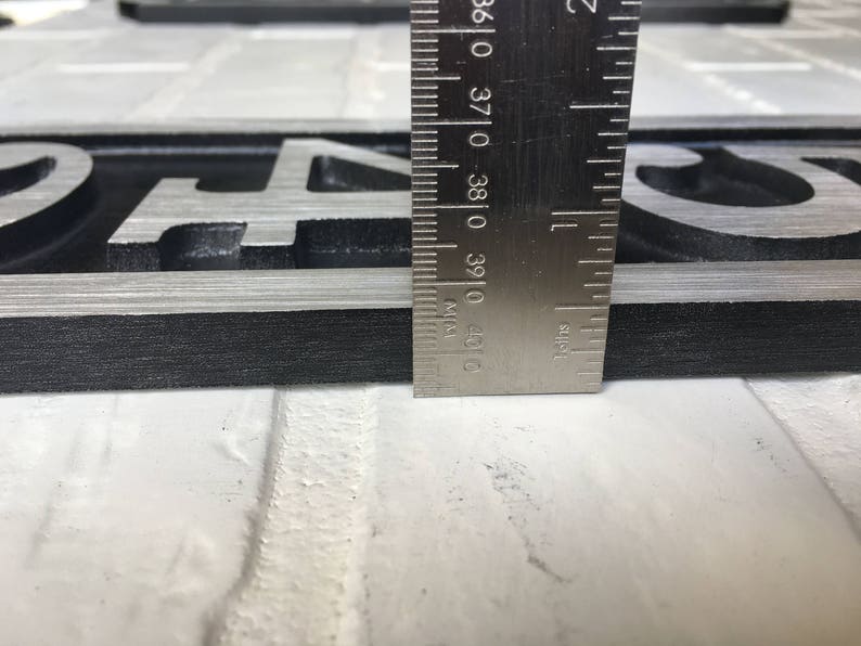 Puede incluir: Primer plano de una placa de n&uacute;mero de casa de metal negro y plateado con el n&uacute;mero "4" en ella. Una regla se coloca junto a la placa para mostrar el tama&ntilde;o. La regla est&aacute; marcada en pulgadas.