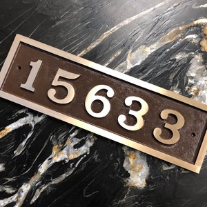 May include: A rectangular house number plaque with a brown background and silver raised numbers. The numbers are 15633.