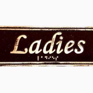 May include: A brown rectangular sign with gold lettering that reads "Ladies" with braille underneath.