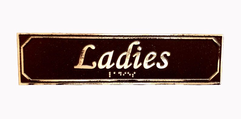 May include: A brown rectangular sign with gold lettering that reads "Ladies" with braille underneath.