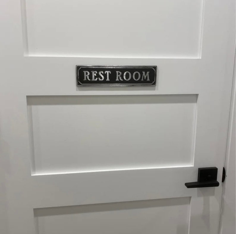 Pu&ograve; includere: Una porta bianca con una maniglia nera e un cartello nero con la scritta "REST ROOM" in lettere argentate.