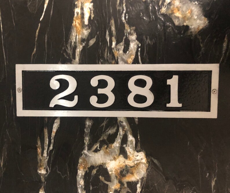 May include: Rectangular black address plaque with the numbers "2381" in a silver font. The plaque has a silver border and is mounted on a black and gold marble-like surface. Two screws secure the plaque.