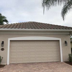May include: A beige house with a brown tiled roof and a white garage door. The house has white trim and a white arched doorway above the garage door. The house number 1271 is visible on the side of the house.