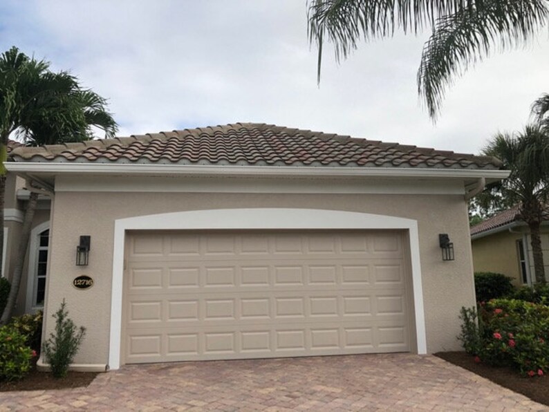May include: A beige house with a brown tiled roof and a white garage door. The house has white trim and a white arched doorway above the garage door. The house number 1271 is visible on the side of the house.