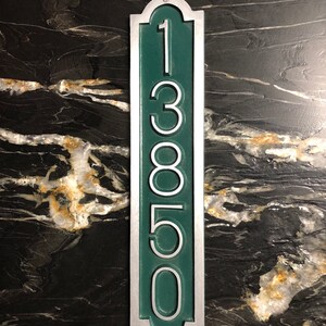 May include: A vertical house number sign with a silver frame and a green background. The number 13850 is displayed in white on the sign.