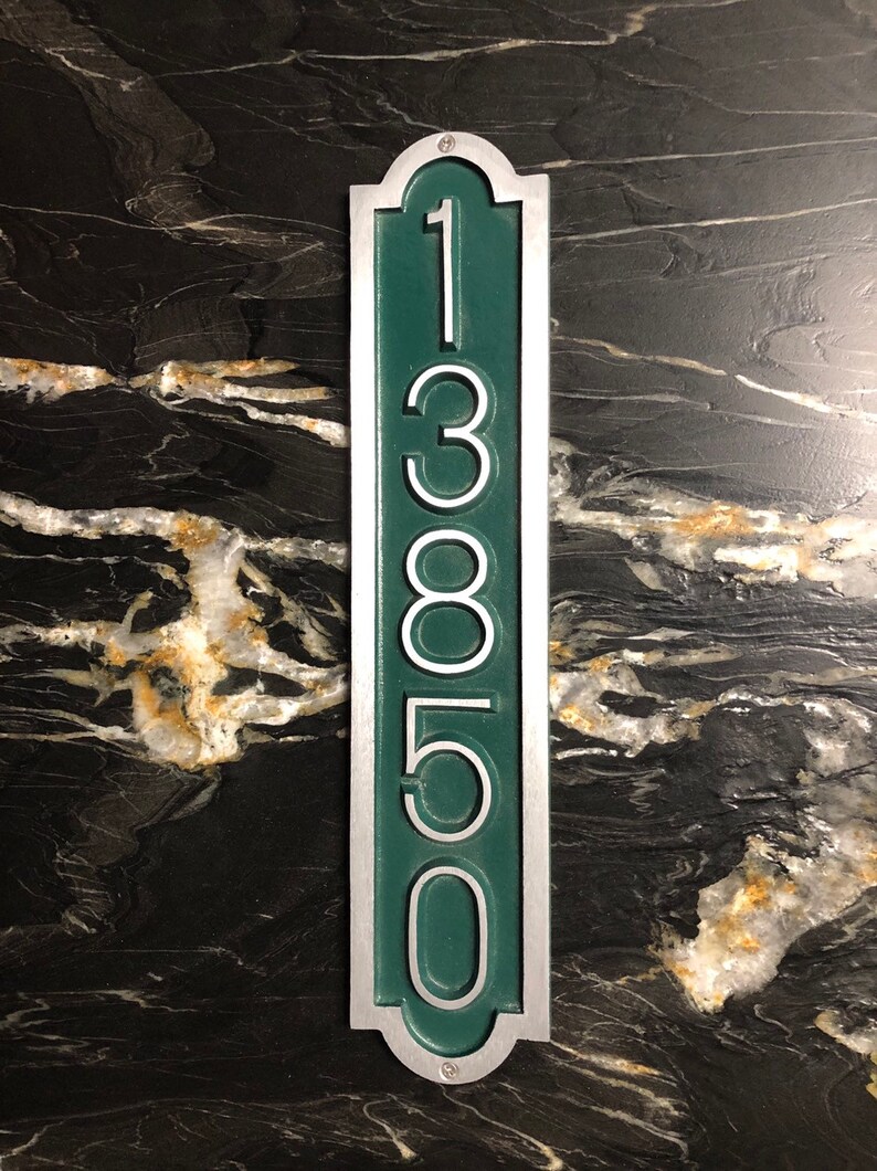 May include: A vertical house number sign with a silver frame and a green background. The number 13850 is displayed in white on the sign.