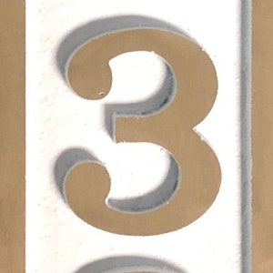 May include: A white background with a gold number three. The number is raised and has a shadow.