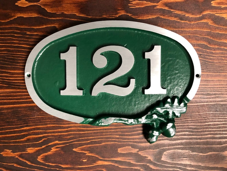 May include: Oval-shaped house number sign with a green background and silver numbers. The numbers "121" are displayed in a bold, sans-serif font. The sign has a silver border and a decorative acorn and leaf design at the bottom.