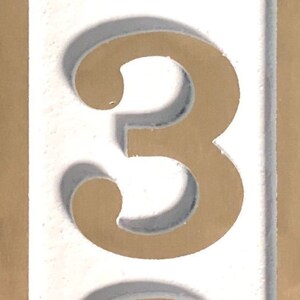 May include: A white background with a gold number three. The number is raised and has a shadow.