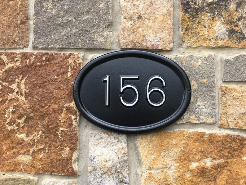 May include: Black oval-shaped house number plaque with white numbers "156" on a stone wall.
