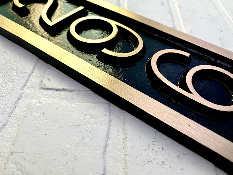 May include: A gold-colored metal sign with the word "CO" in raised lettering on a black background. The sign has a brushed gold border.