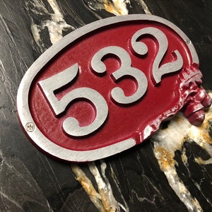 May include: A red oval-shaped house number sign with silver lettering. The number 532 is displayed on the sign. The sign is slightly damaged on the right side.
