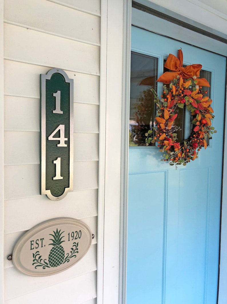 May include: A white house with a blue door and a green and silver house number plaque that says "141". There is a fall wreath on the door with orange and red leaves and a brown bow. There is a white oval sign with a green pineapple and the text "EST. 1920".