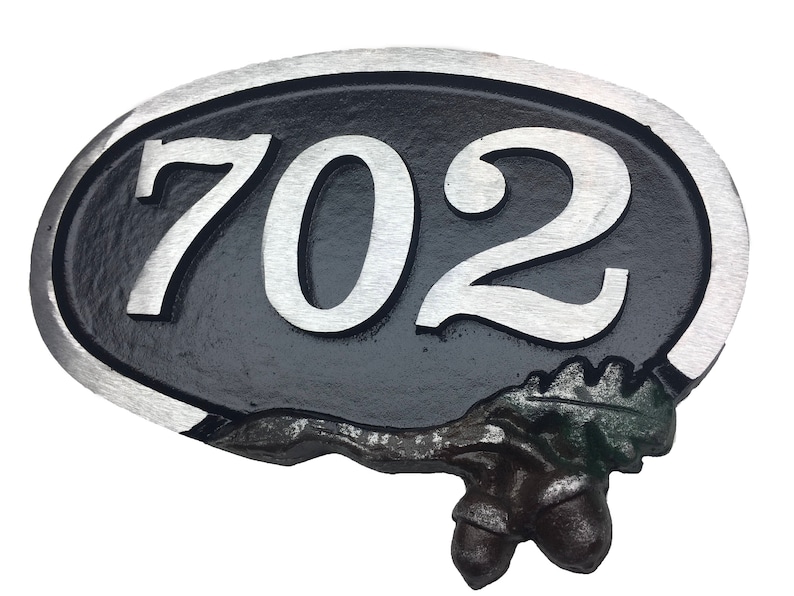 May include: A black oval-shaped house number sign with a silver border and silver numbers "702". The sign has a decorative acorn and leaf design at the bottom.
