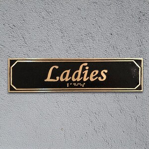 May include: A black rectangular sign with gold trim and rounded corners. The sign reads "Ladies" in gold cursive lettering with braille below.