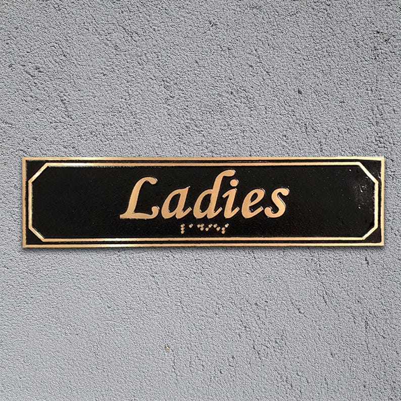 May include: A black rectangular sign with gold trim and rounded corners. The sign reads "Ladies" in gold cursive lettering with braille below.