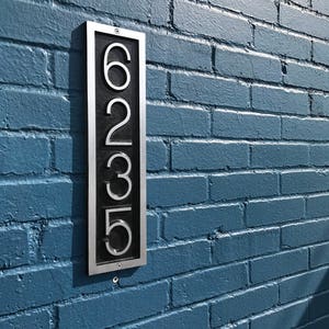 May include: A silver metal house number plaque with black numbers reading "6235" mounted on a blue brick wall.