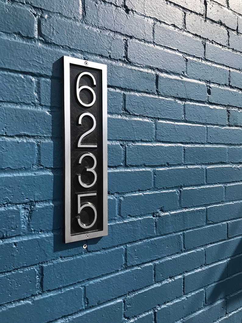 May include: A silver metal house number plaque with black numbers reading "6235" mounted on a blue brick wall.